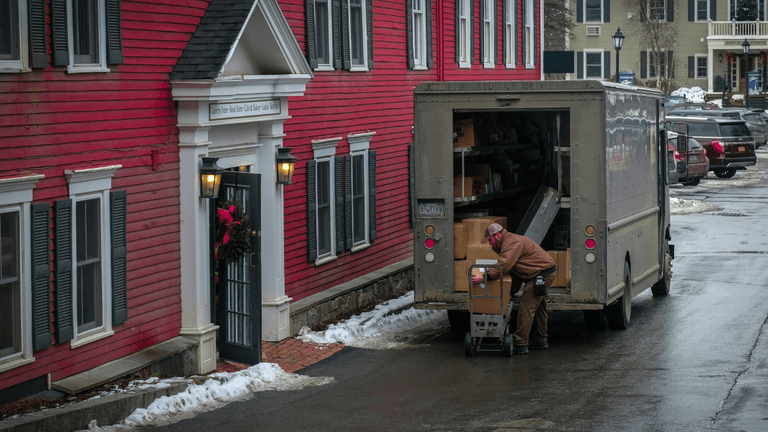 ChatGPT said: This image shows a delivery driver unloading packages from the back of a truck near a red building decorated with a wreath. The driver is using a dolly to transport a large box toward the building. The scene suggests a residential or small business delivery, with snow on the ground and cars parked nearby. The front of the building has a sign indicating it might be a store or office, with the name visible above the door.