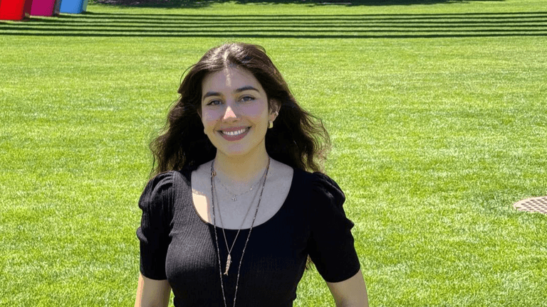 "Anna Belhassen smiling outdoors, standing in front of colorful rainbow sculptures, showcasing her work in engineering and design."