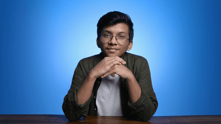 Portrait of a young man with glasses sitting at a table against a blue background.