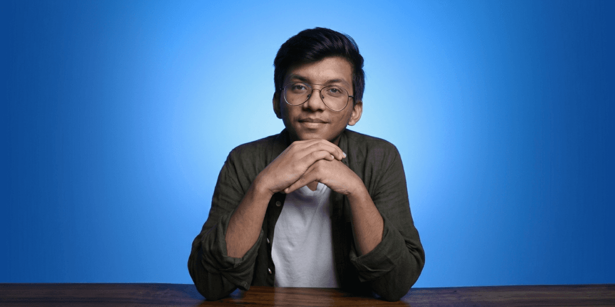 Portrait of a young man with glasses sitting at a table against a blue background.