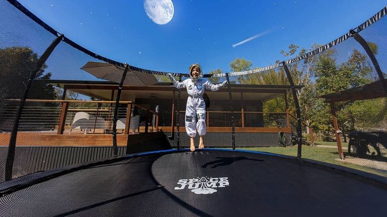 Child in astronaut suit jumping on SpaceJump trampoline with moon backdrop in backyard.