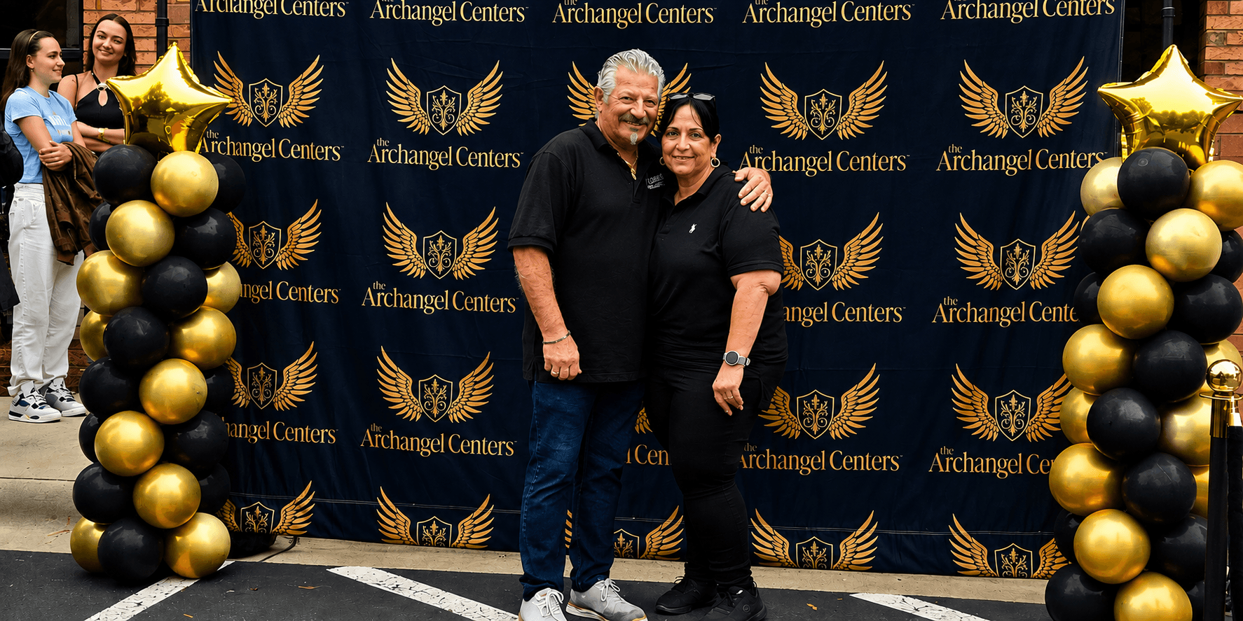 Francesco and Carmela Ventura at an Archangel Centers event with branded backdrop and black-and-gold balloon display.