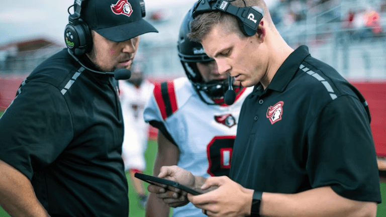 Coaches wearing headsets review a tablet with a football player on the sideline.