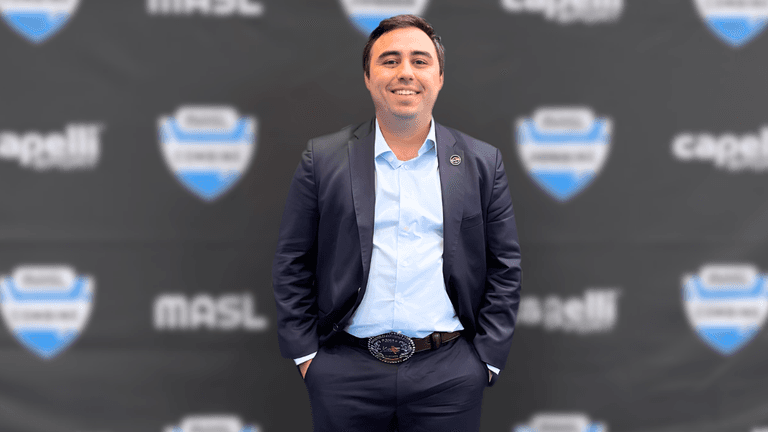 Smiling man in suit standing before branded backdrop, hands in pockets, professional event setting.