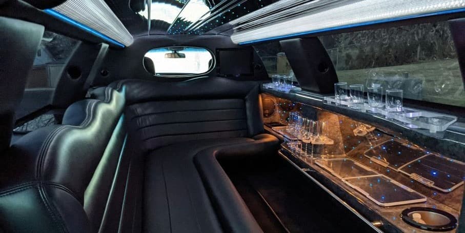 Interior of a luxury limousine featuring sleek leather seating, ambient lighting, and glassware along the bar area.