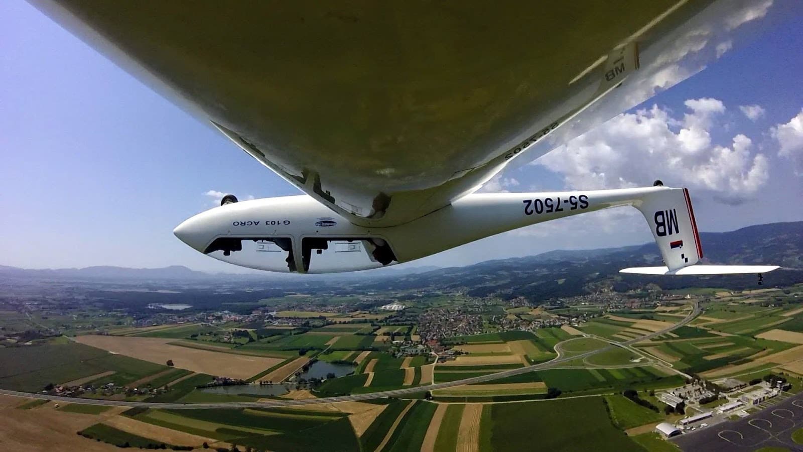 A glider soaring over lush fields with stunning mountain views below.