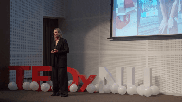 Dr. Irina Zelenkova delivering a TEDx Talk on high performance and altitude training, standing beside a large 'TEDx' sign, while showcasing her scientific approach to achieving extraordinary feats.