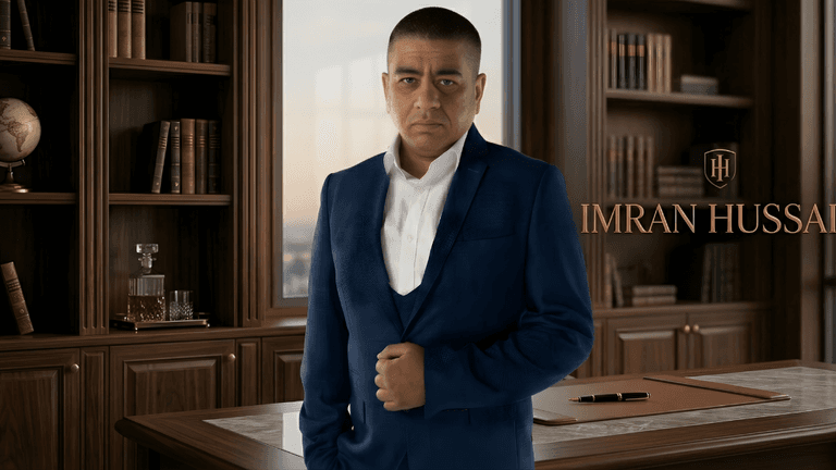 Man in navy suit standing in office with bookshelves, labeled Imran Hussain.
