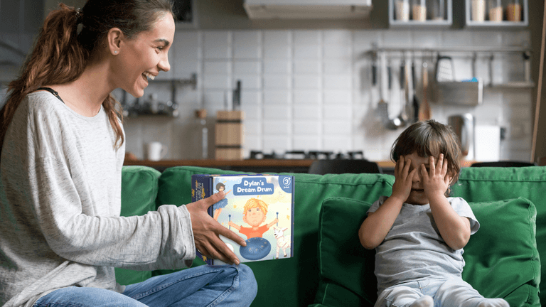 A mother sitting on a green couch, smiling and holding the 'Dylan's Dream Drum' box, while her young son playfully covers his eyes with his hands. The kitchen is visible in the background with modern appliances.