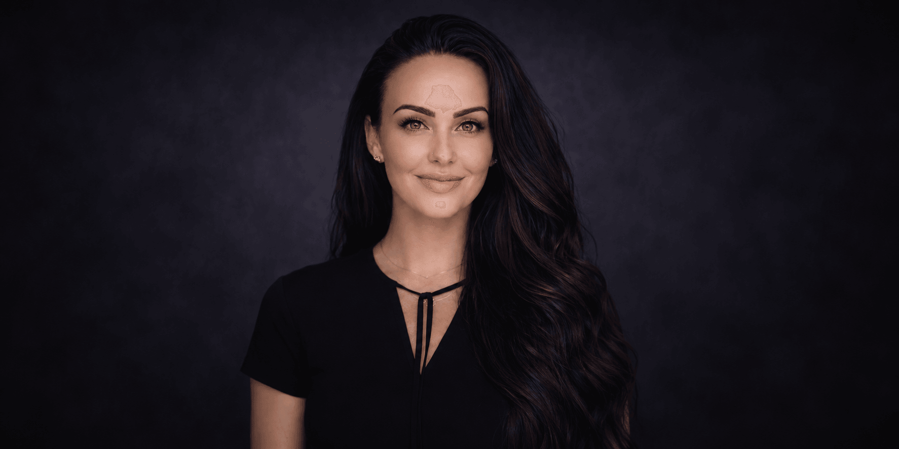 A woman with long dark hair and blue eyes smiles against a dark background