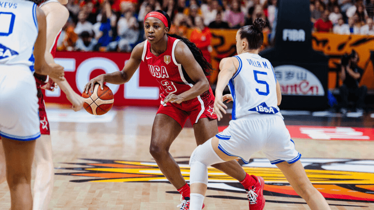 Basketball game with a player in red dribbling while two defenders approach.