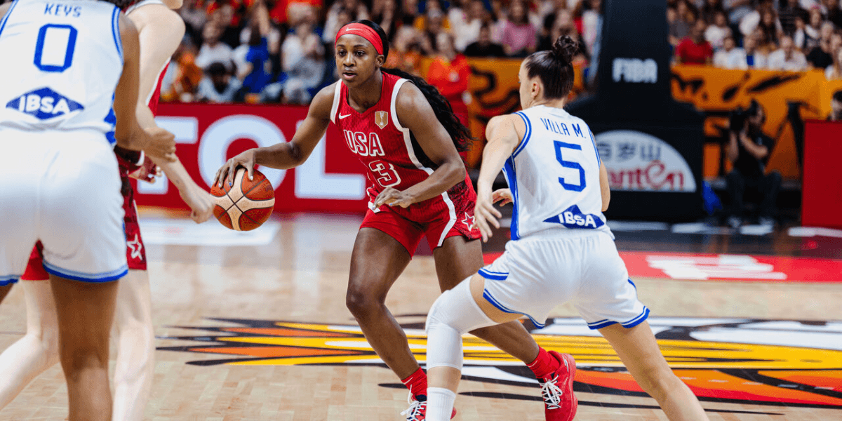 Basketball game with a player in red dribbling while two defenders approach.
