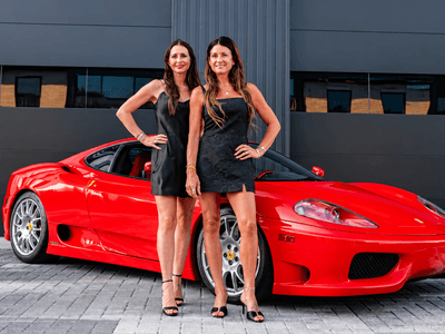 Two women in black dresses pose in front of a red sports car.