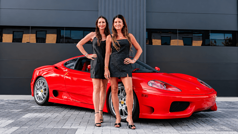 Two women in black dresses pose in front of a red sports car.