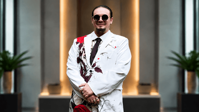 Man in a white suit with red floral patterns and sunglasses, standing confidently indoors