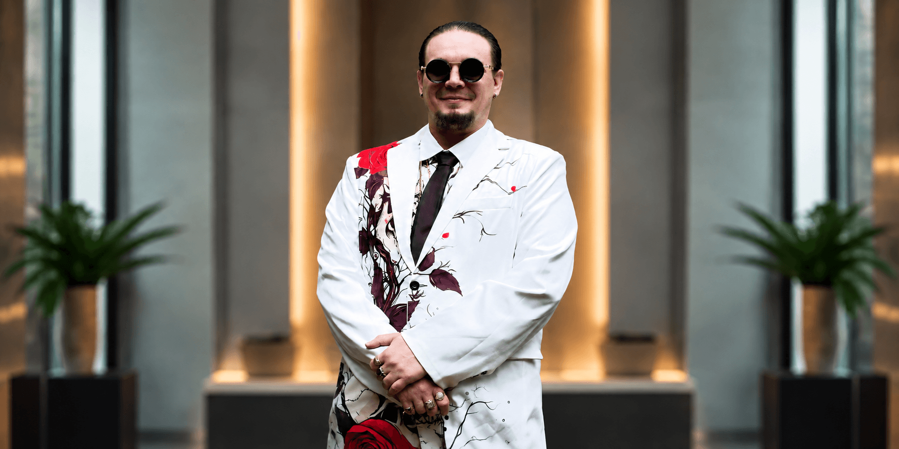 Man in a white suit with red floral patterns and sunglasses, standing confidently indoors