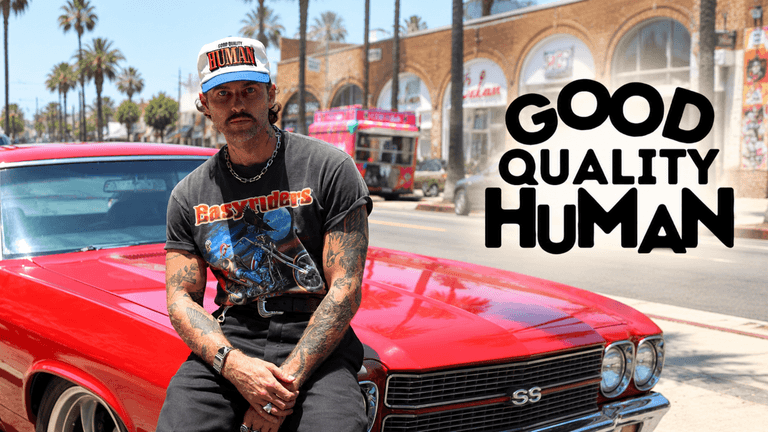 A tattooed man in a vintage t-shirt sits on a red car outdoors