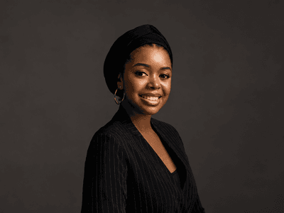 Young woman wearing a black headscarf and pinstripe blazer, smiling against a dark background
