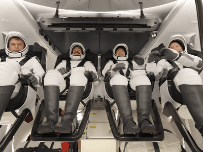 Four astronauts seated inside a spacecraft, wearing space suits and helmets, prepared for launch