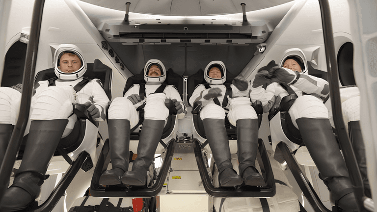 Four astronauts seated inside a spacecraft, wearing space suits and helmets, prepared for launch
