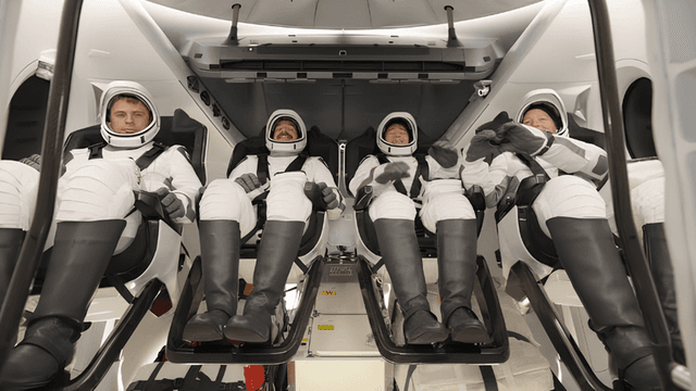Four astronauts seated inside a spacecraft, wearing space suits and helmets, prepared for launch