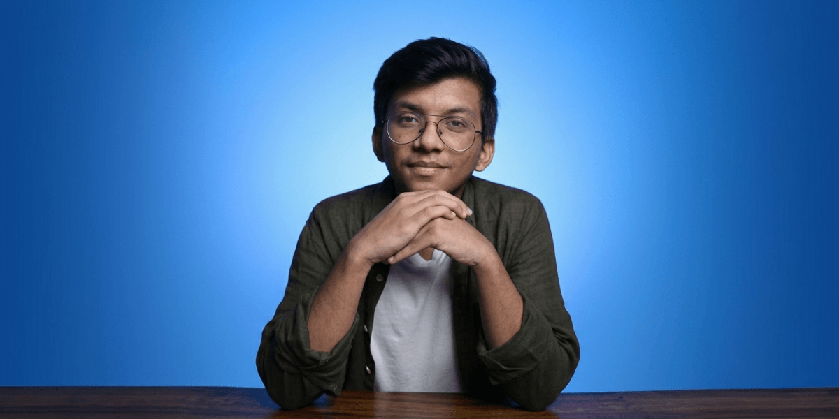 Portrait of a young man with glasses sitting at a table against a blue background.