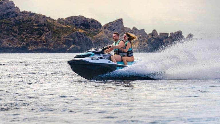 Two riders travel on an Aquamarine jet ski across the Mediterranean with rocky coastline behind them.