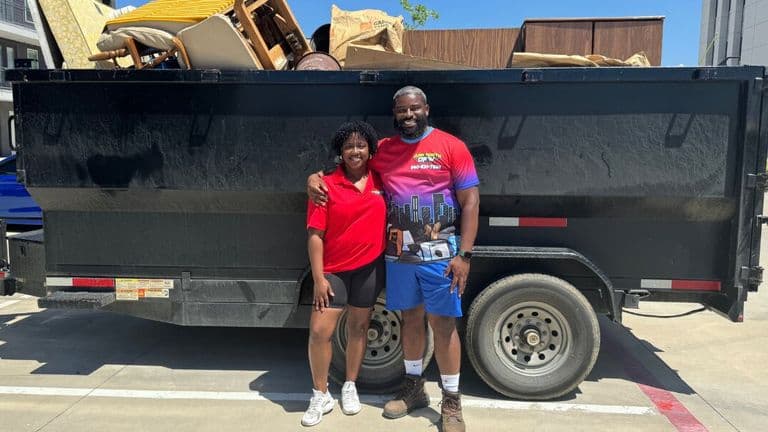 A smiling man and woman stand arm in arm in front of a large black trailer filled with furniture and bags. They appear content and proud.
