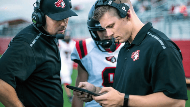 Coaches wearing headsets review a tablet with a football player on the sideline.