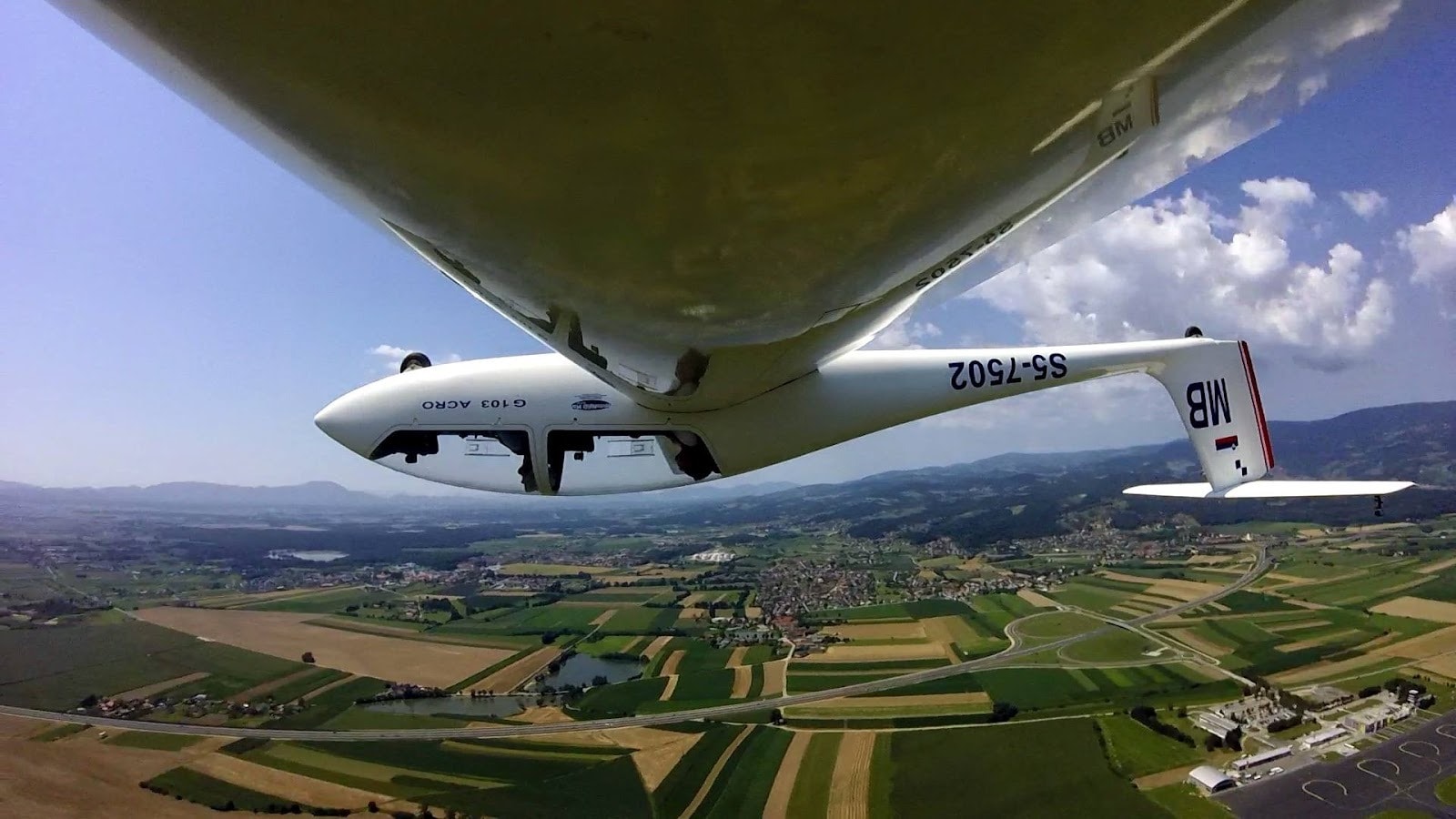 A glider soaring over lush fields with stunning mountain views below.