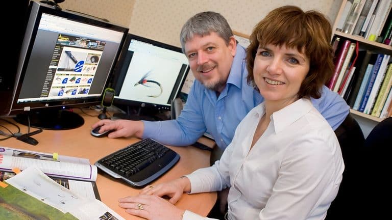 A man and woman collaborate at a desk, focused on computer monitors, discussing fly fishing resources and community building.
