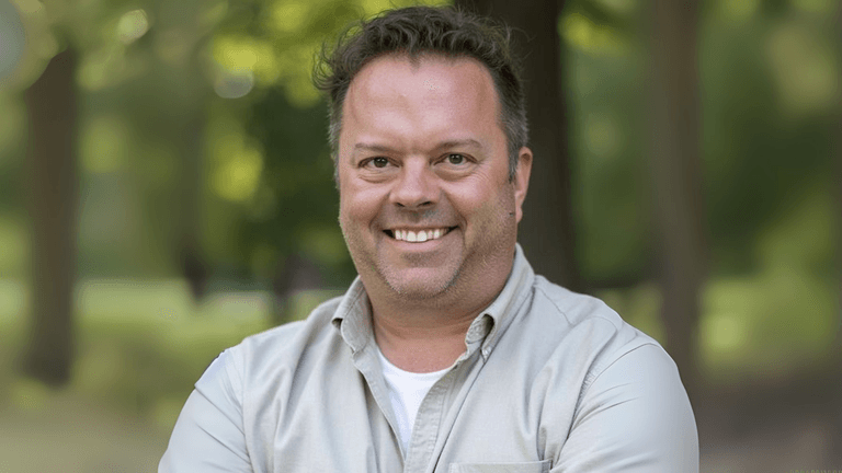 Smiling man in a casual white shirt and khaki jacket standing in a park setting with blurred trees in the background
