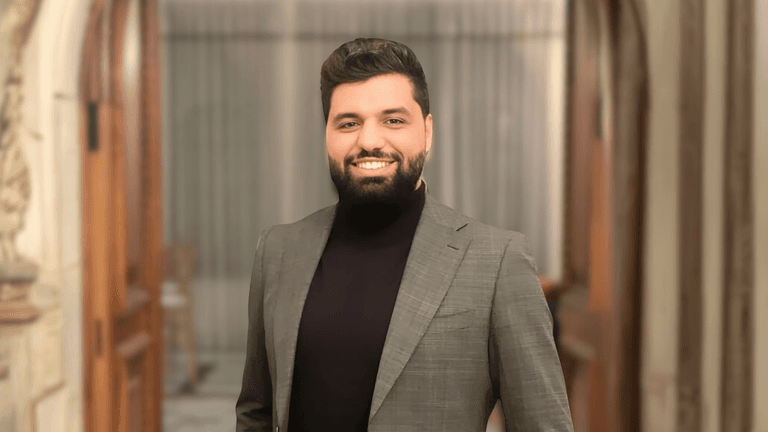 Professional portrait of a man in a suit and turtleneck, smiling confidently in a sophisticated indoor setting with wooden doors and neutral-toned background.