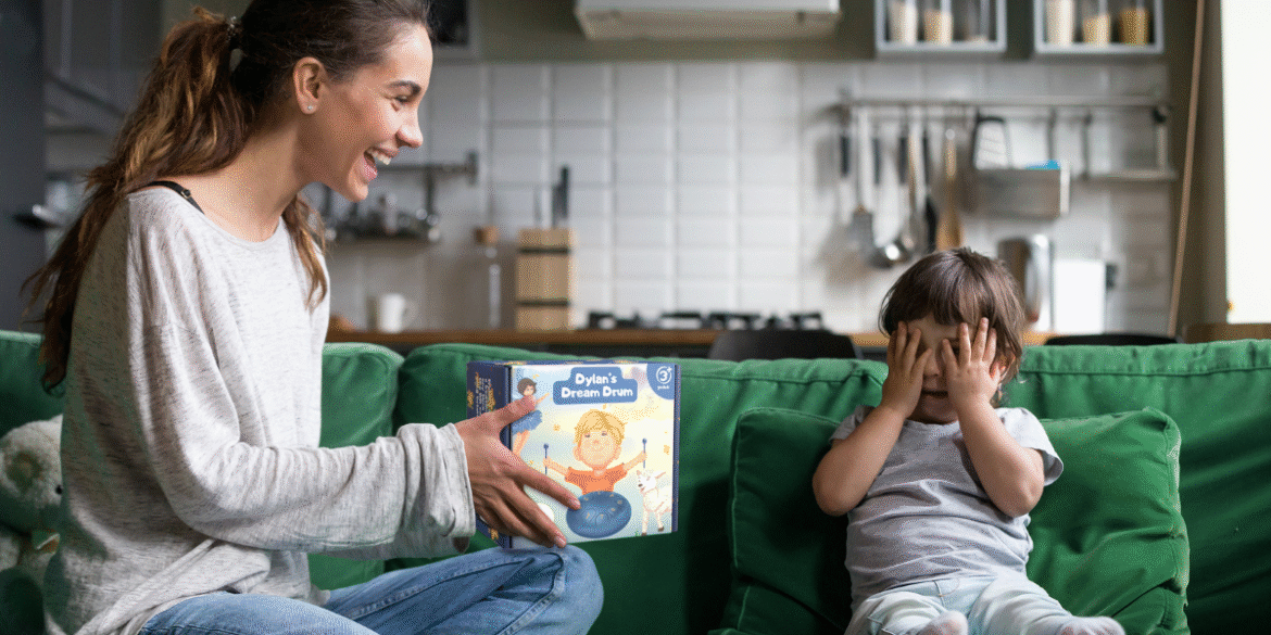 A mother sitting on a green couch, smiling and holding the 'Dylan's Dream Drum' box, while her young son playfully covers his eyes with his hands. The kitchen is visible in the background with modern appliances.