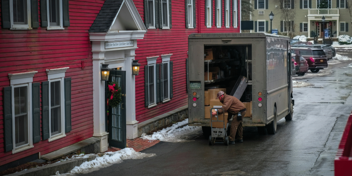ChatGPT said: This image shows a delivery driver unloading packages from the back of a truck near a red building decorated with a wreath. The driver is using a dolly to transport a large box toward the building. The scene suggests a residential or small business delivery, with snow on the ground and cars parked nearby. The front of the building has a sign indicating it might be a store or office, with the name visible above the door.