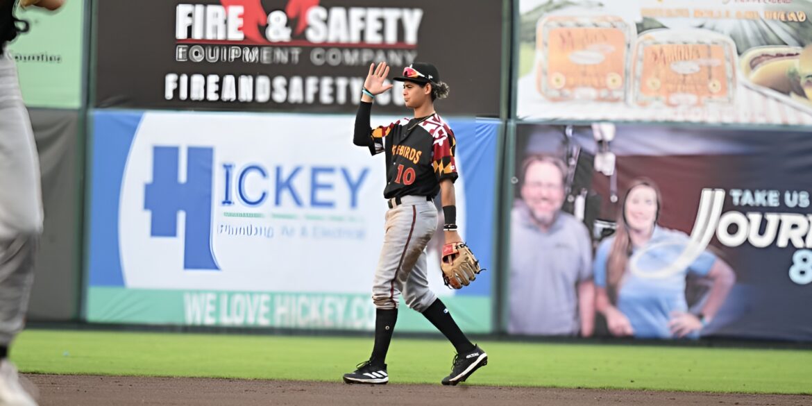 Player in a black and red Shorebirds uniform, walking on the field and waving while holding a glove.