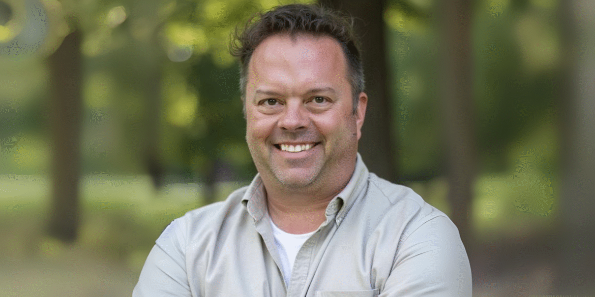 Smiling man in a casual white shirt and khaki jacket standing in a park setting with blurred trees in the background