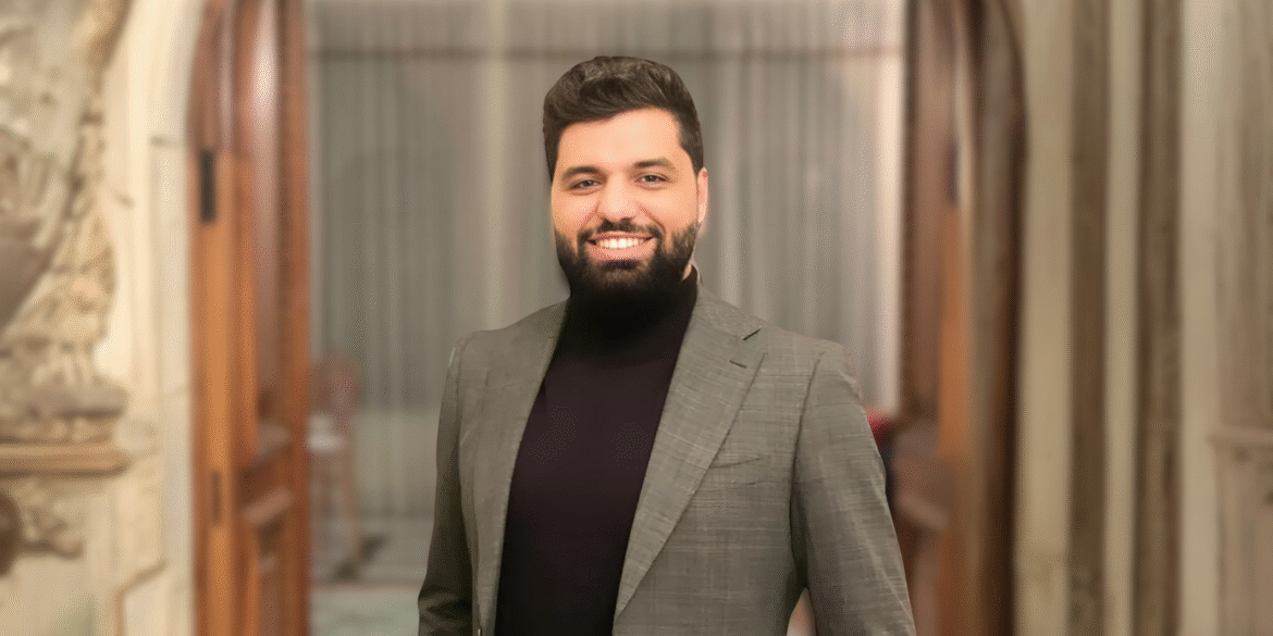 Professional portrait of a man in a suit and turtleneck, smiling confidently in a sophisticated indoor setting with wooden doors and neutral-toned background.