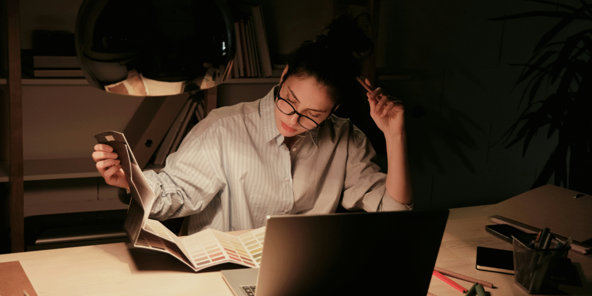Woman working late at desk, reviewing color swatches, symbolizing stress and decision-making in a high-pressure environment.