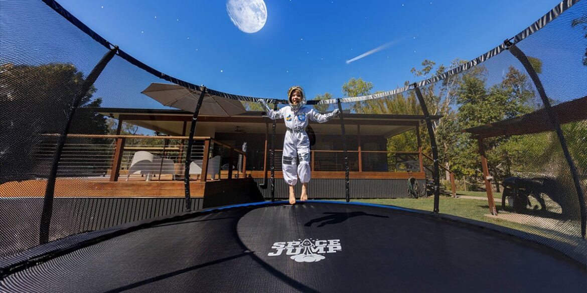 Child in astronaut suit jumping on SpaceJump trampoline with moon backdrop in backyard.