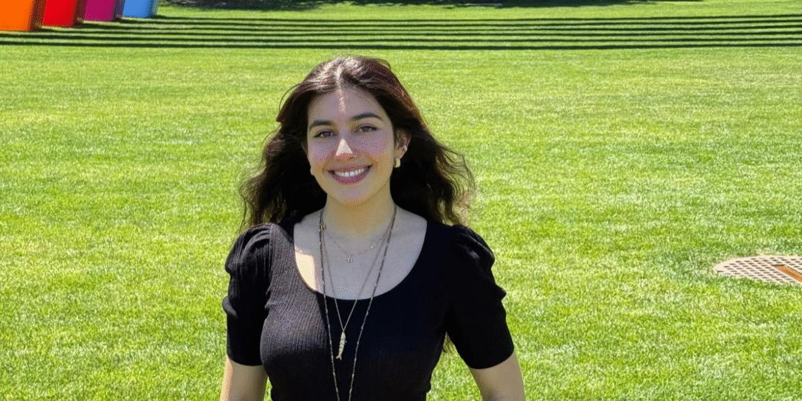 "Anna Belhassen smiling outdoors, standing in front of colorful rainbow sculptures, showcasing her work in engineering and design."