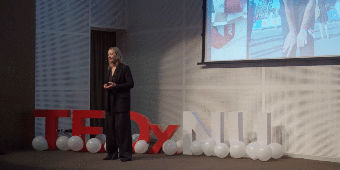 Dr. Irina Zelenkova delivering a TEDx Talk on high performance and altitude training, standing beside a large 'TEDx' sign, while showcasing her scientific approach to achieving extraordinary feats.