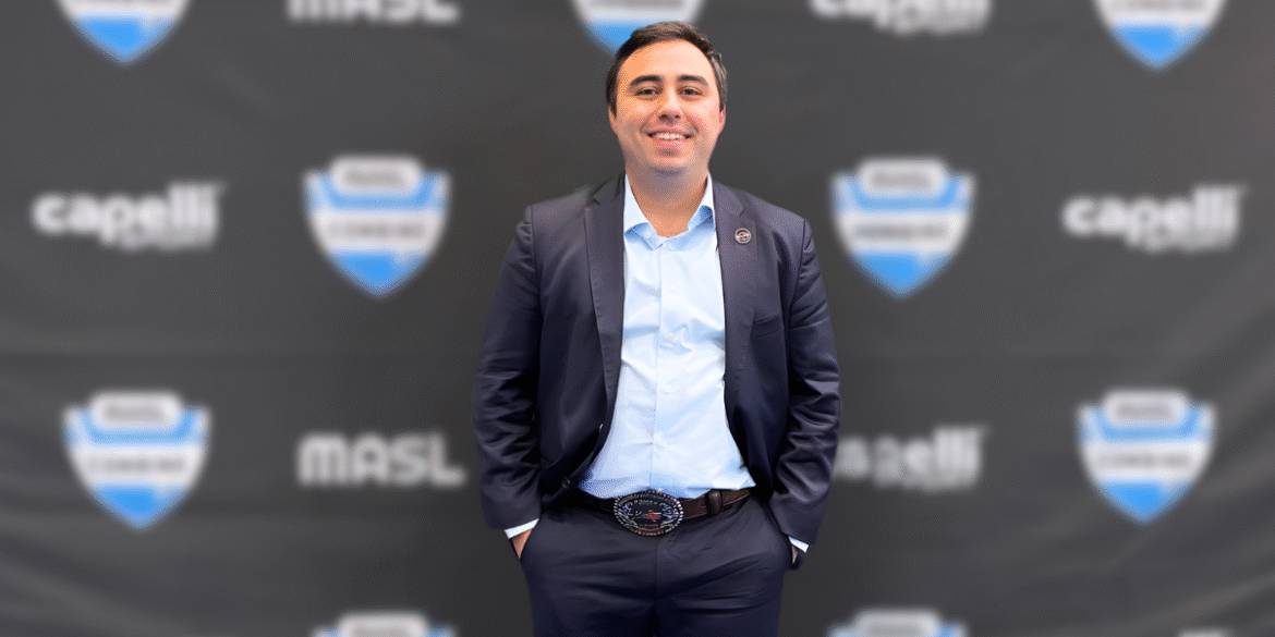 Smiling man in suit standing before branded backdrop, hands in pockets, professional event setting.