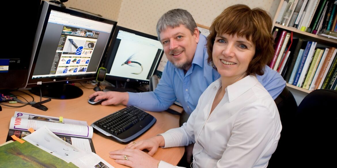 A man and woman collaborate at a desk, focused on computer monitors, discussing fly fishing resources and community building.