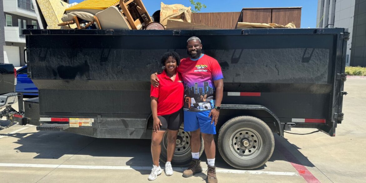 A smiling man and woman stand arm in arm in front of a large black trailer filled with furniture and bags. They appear content and proud.
