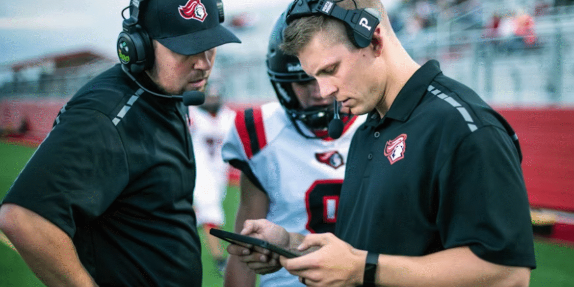 Coaches wearing headsets review a tablet with a football player on the sideline.