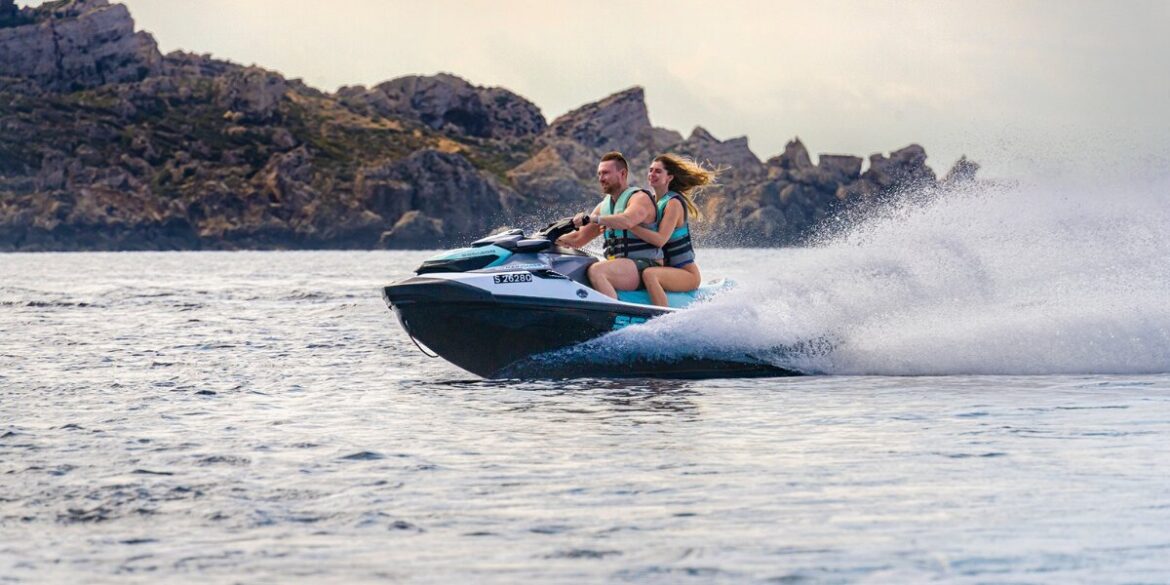 Two riders travel on an Aquamarine jet ski across the Mediterranean with rocky coastline behind them.